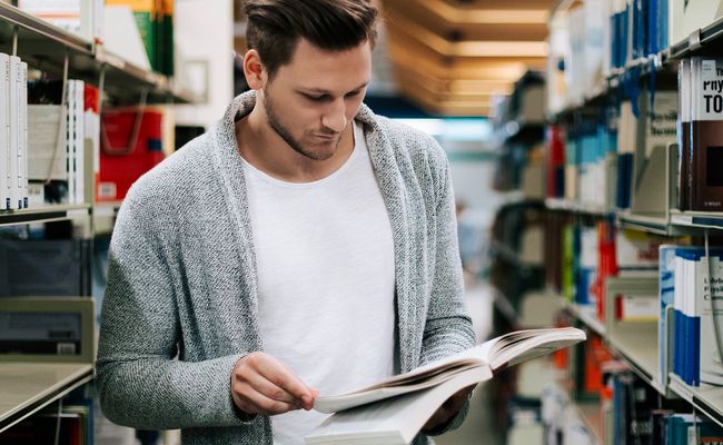 lesender Studierender zwischen zwei Bücherregalen stehend
